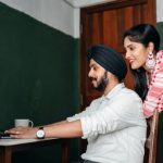 Home Office - Side view of positive young Sikh man in shirt and turban working on laptop at home while wife leaning on chair behind