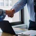 Laptop Handshake - Crop unrecognizable coworkers in formal wear standing at table with laptop and documents while greeting each other before meeting