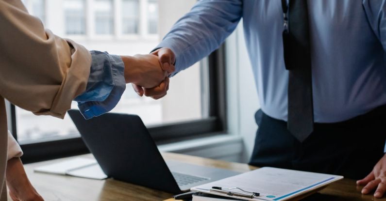 Laptop Handshake - Crop unrecognizable coworkers in formal wear standing at table with laptop and documents while greeting each other before meeting