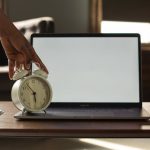 Clock Plan - Anonymous black person touching alarm clock near opened netbook with white blank screen placed on table with notebook and pens