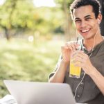 Laptop Sunshine - Cheerful guy with laptop and earphones sitting in park while drinking juice and smiling at camera