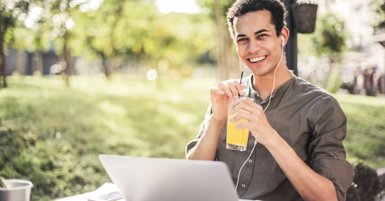 Laptop Sunshine - Cheerful guy with laptop and earphones sitting in park while drinking juice and smiling at camera