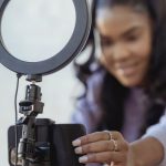 Social Media Phone - Cheerful young African American female blogger in stylish sweater smiling while setting up camera of smartphone attached to tripod with ring light before recording vlog