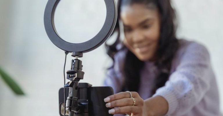 Social Media Phone - Cheerful young African American female blogger in stylish sweater smiling while setting up camera of smartphone attached to tripod with ring light before recording vlog