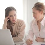 Remote Internship - Pensive female entrepreneurs wearing casual clothes sitting together at table with laptop and talking about problems in project