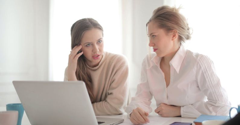 Remote Internship - Pensive female entrepreneurs wearing casual clothes sitting together at table with laptop and talking about problems in project