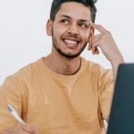 Future Work - Smiling young bearded Hispanic male entrepreneur thinking over new ideas for startup project and looking away dreamily while working at table with laptop and taking notes in notebook