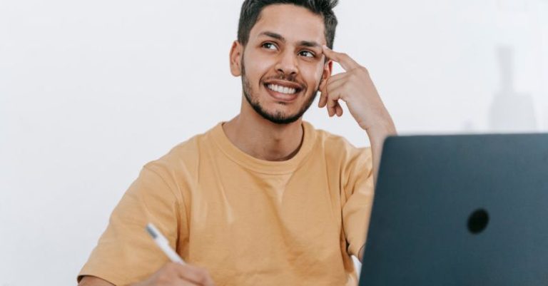 Future Work - Smiling young bearded Hispanic male entrepreneur thinking over new ideas for startup project and looking away dreamily while working at table with laptop and taking notes in notebook
