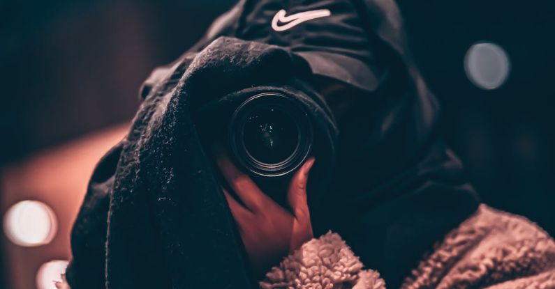 Hidden Job - Unrecognizable person wearing hooded clothes and hiding face behind black scarf taking photo on professional camera on blurred background