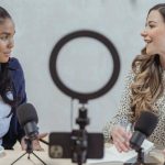 Phone Interview - Positive blogger asking questions to African American speaker and recording interview on cellphone at table with microphones