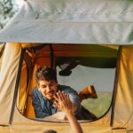 Ladder Success - Back view of unrecognizable young female camper standing on ladder and giving high five to smiling boyfriend recreating in tent placed on SUV car roof at lakeside