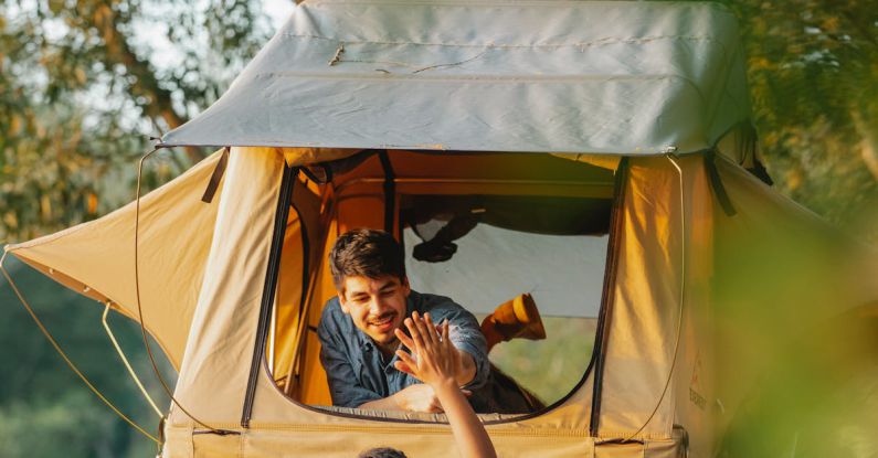 Ladder Success - Back view of unrecognizable young female camper standing on ladder and giving high five to smiling boyfriend recreating in tent placed on SUV car roof at lakeside