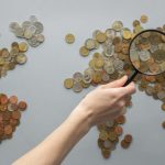 Paycheck Magnifier - Top view of crop unrecognizable traveler with magnifying glass standing over world map made of various coins on gray background