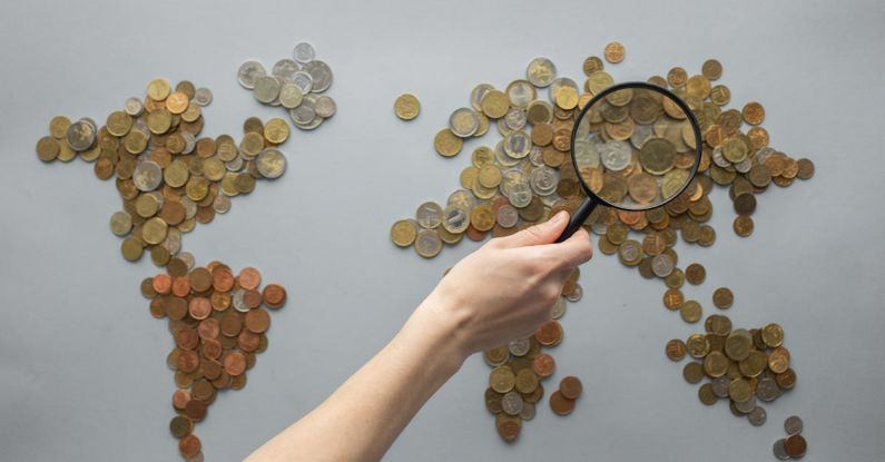 Paycheck Magnifier - Top view of crop unrecognizable traveler with magnifying glass standing over world map made of various coins on gray background